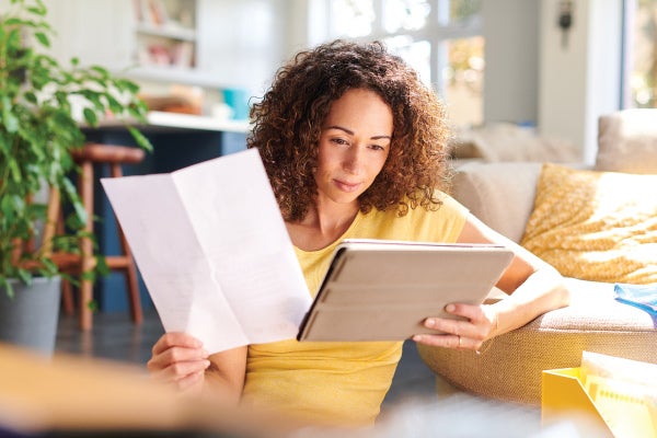 Woman in yellow shirt, reading a tablet and holding a paper, seated on a cozy couch.