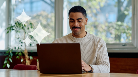 Man smiling while using a laptop in a bright, plant-filled room.