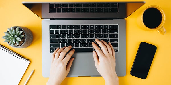 Hands typing on a laptop, yellow desk, coffee, plant, notebook, phone nearby.