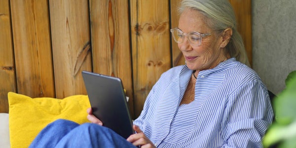 Elderly woman reading a tablet, seated by a wooden wall with a yellow pillow.