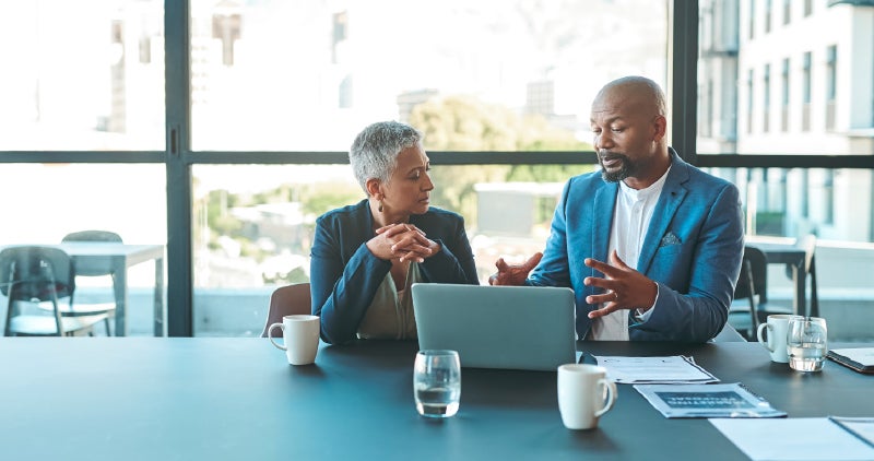 Two colleagues discussing at a table with a laptop and documents.
